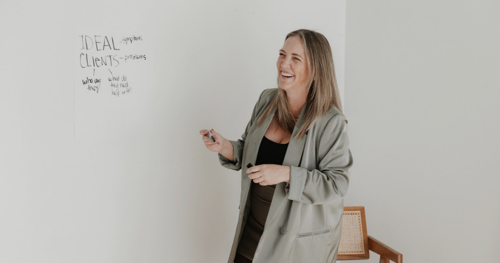 girl standing laughing holding a pen with a poster on the wall that says ideal clients 